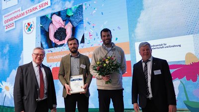 Vier Personen stehen auf einer Bühne, eine hält eine Urkunde und eine andere einen Blumenstrauß in der Hand