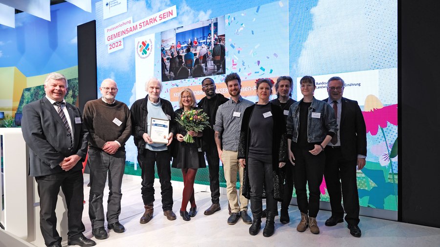 Zehn Personen stehen auf einer Bühne, eine hält eine Urkunde und eine andere einen Blumenstrauß in der Hand, alle lächeln in die Kamera