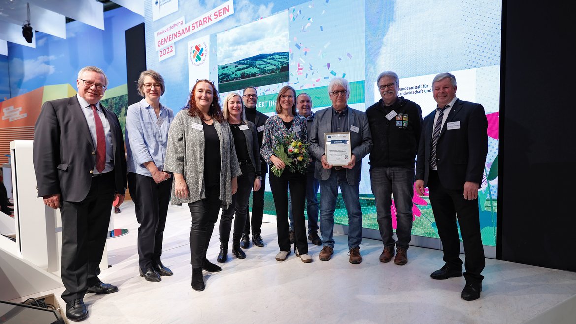 Zehn Personen stehen auf einer Bühne, eine hält eine Urkunde und eine andere einen Blumenstrauß in der Hand, alle lächeln in die Kamera